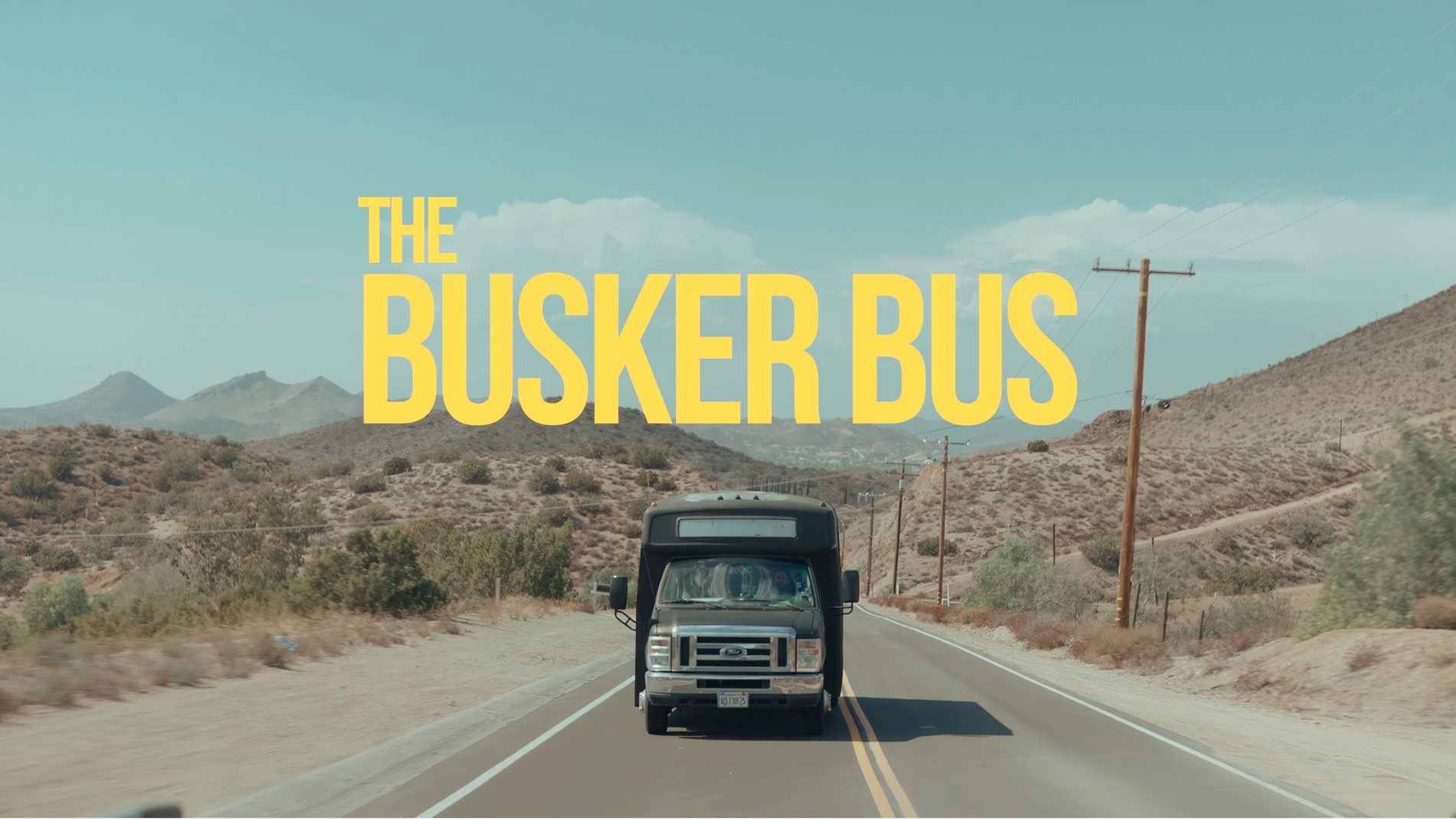 The Busker Bus, a white shuttle bus, driving toward the camera on a desert highway under the yellow title The Busker Bus